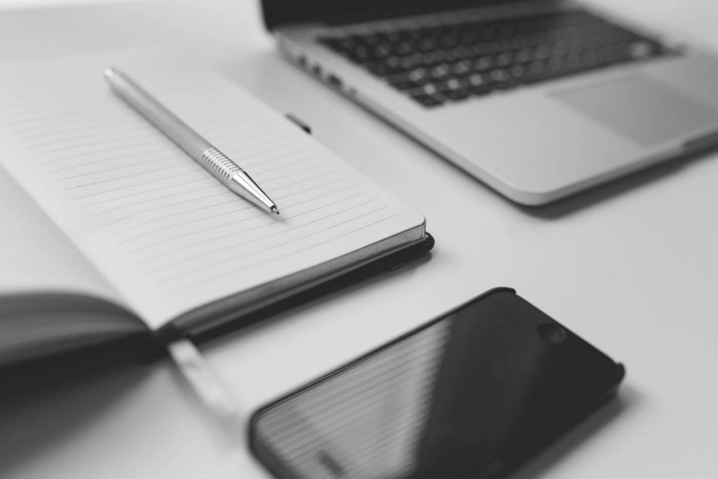 Laptop on desk with a cellphone, and a pen on an open notebook