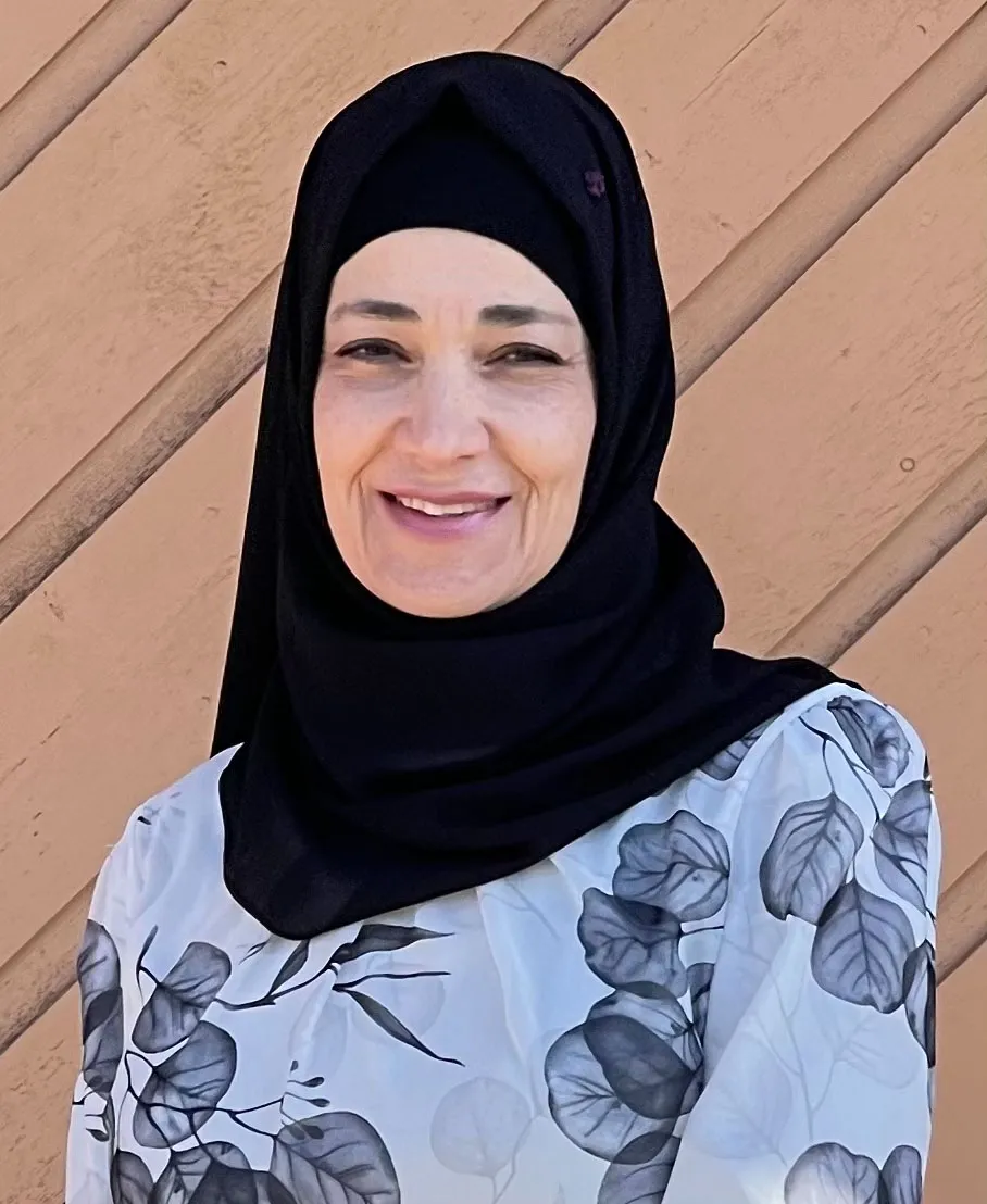A woman with a black headscarf and floral top smiles warmly at the camera.