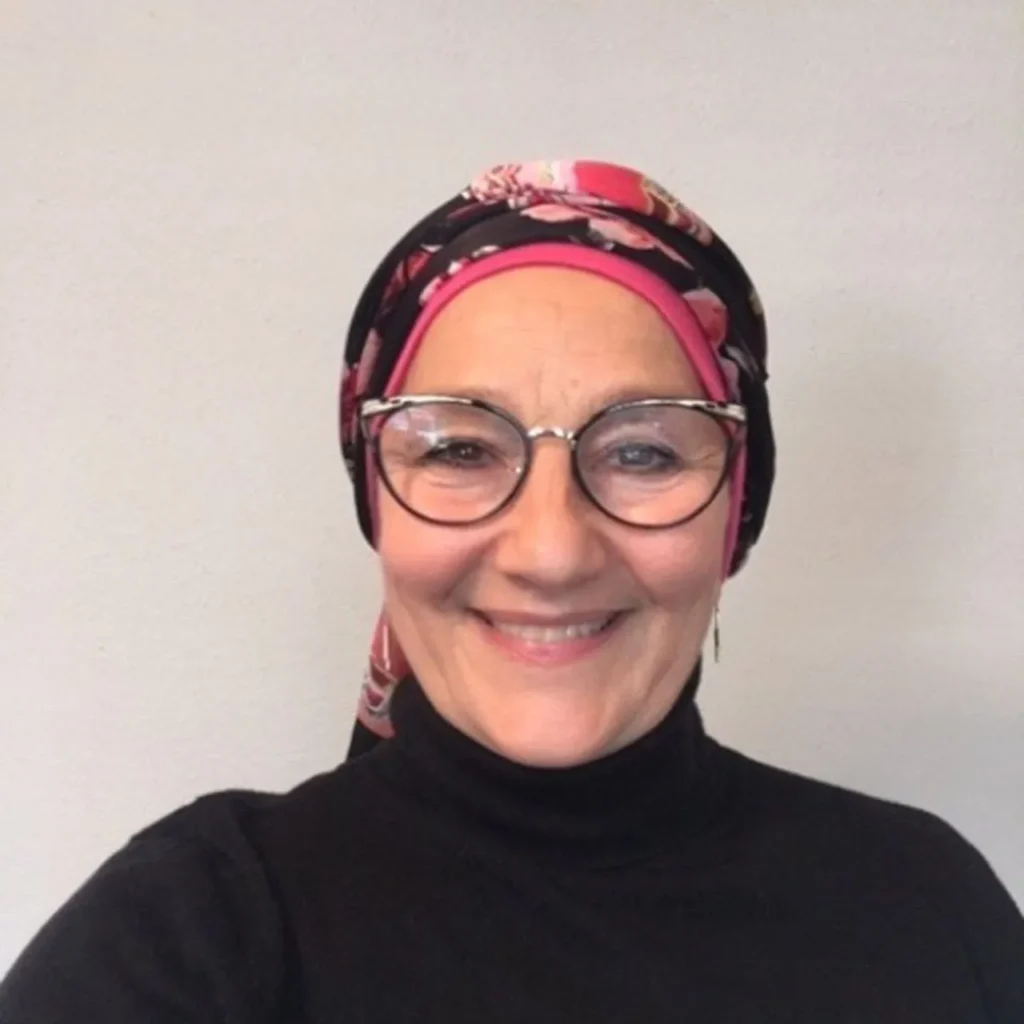 A white woman in eyeglasses, floral headscarf and black high neck smiles warmly at teh camera.